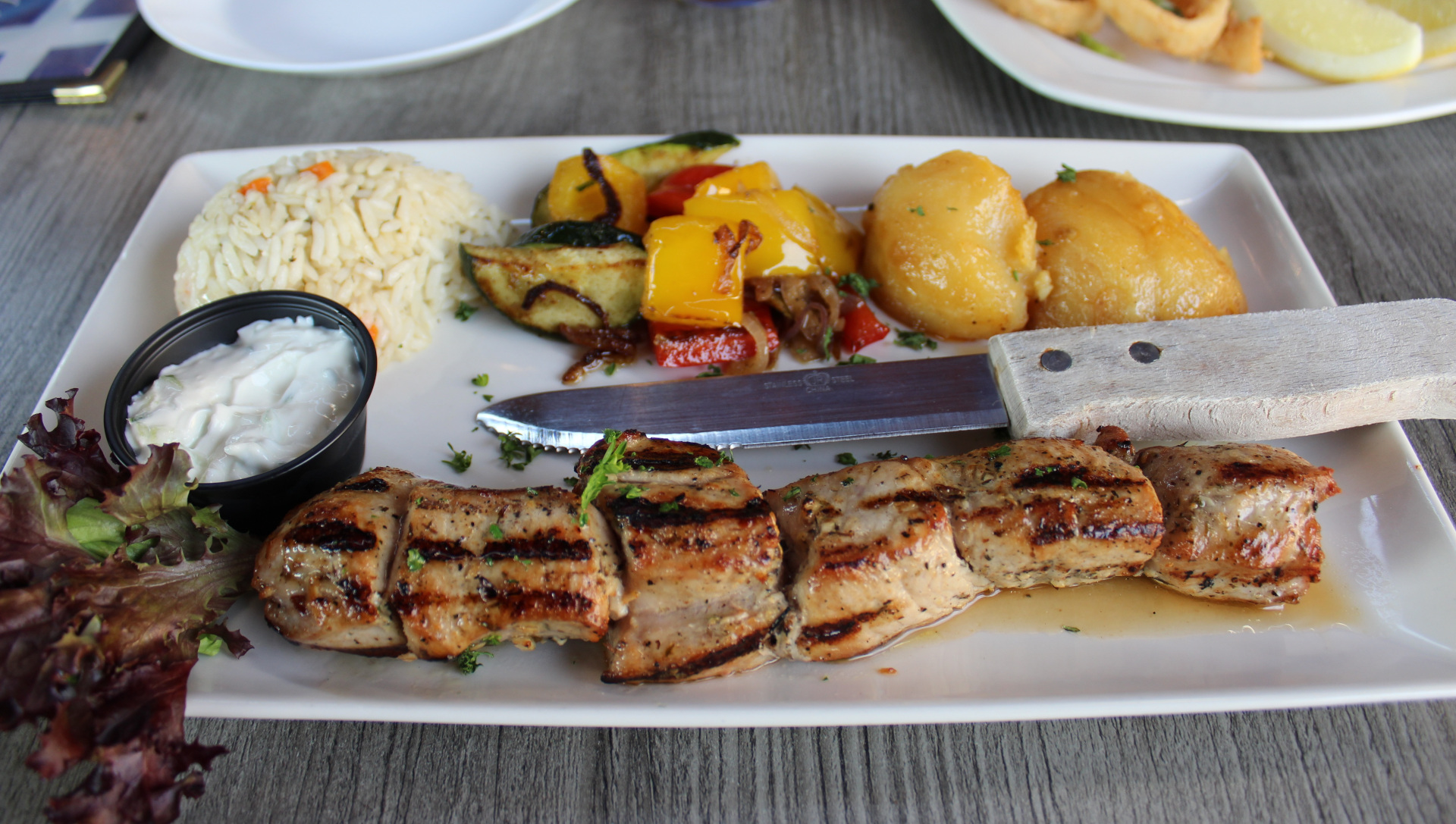 Gegrillte Fleischspieße mit Reis, gebratenem Gemüse, Kartoffeln und Tzatziki auf einem weißen Teller, daneben ein Messer.